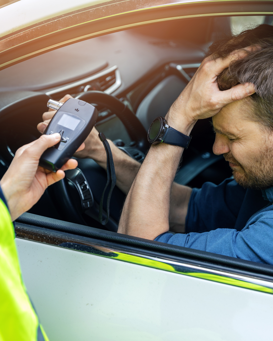 Ein Mann sitzt im Auto und hält sich den Kopf, während ein Polizist ihm ein Atemalkoholtestgerät reicht. Der Mann wirkt besorgt und bedrückt.