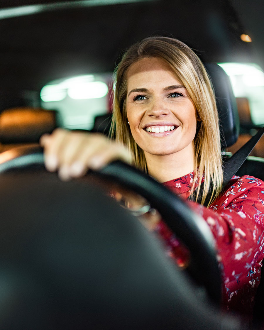 Lächelnde Frau hinter dem Steuer eines Autos, die entspannt nach vorne schaut, mit angeschnalltem Sicherheitsgurt.