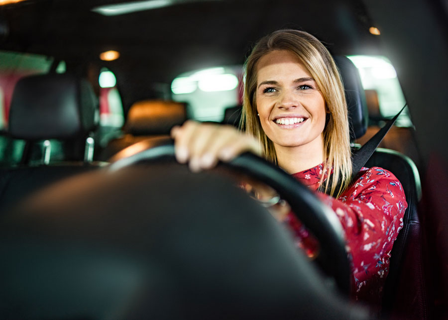 Lächelnde Frau hinter dem Steuer eines Autos, die entspannt nach vorne schaut, mit angeschnalltem Sicherheitsgurt.